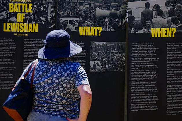 A woman stands in front of a large poster. The poster reads 'Battle of Lewisham' 'What 'When' in yellow text with descriptions below.