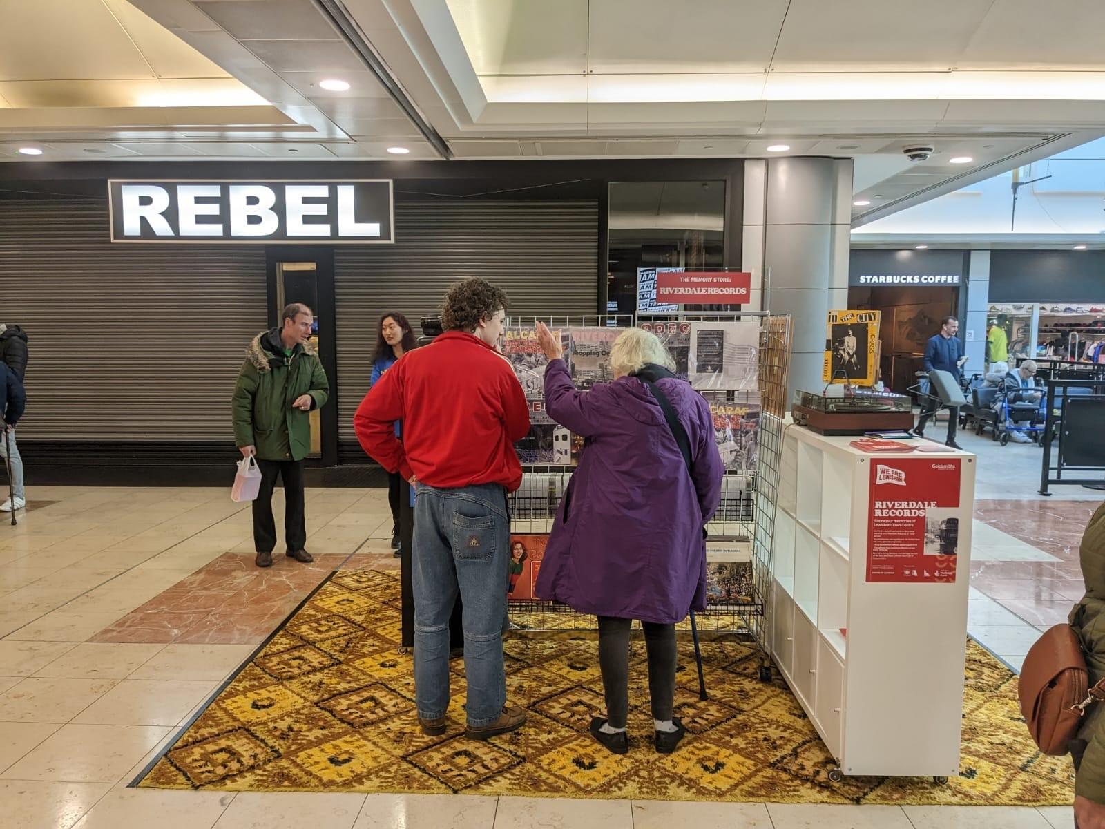A young man in a red jacket stands to the left of an elderly woman. The pair are looking at and discussing an installation of a record store.