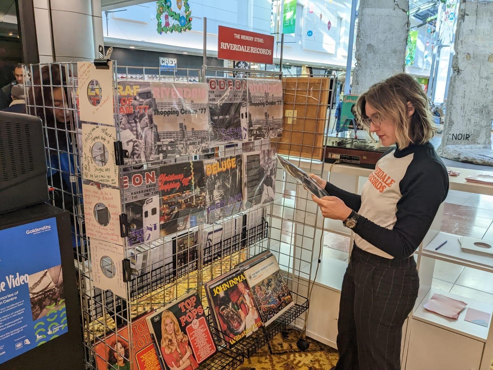 A woman explores an installation of a record store. She is holding and reading the back of a record.